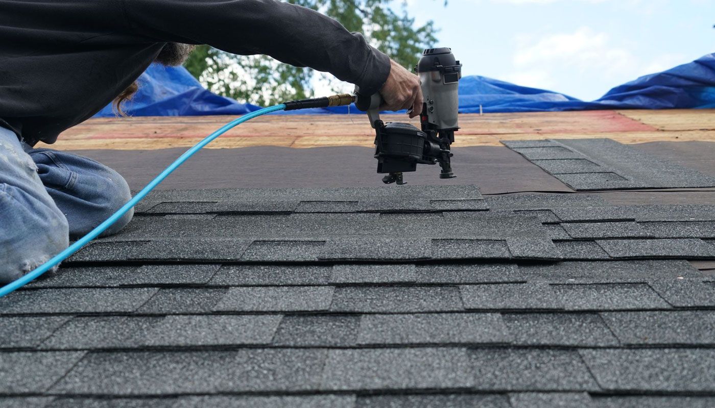 Worker using a nail gun to install roof shingles on a house under blue tarps