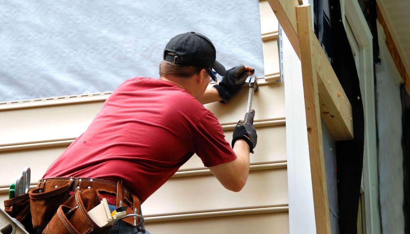 Worker in red shirt installing beige siding on a house exterior with a nail gun