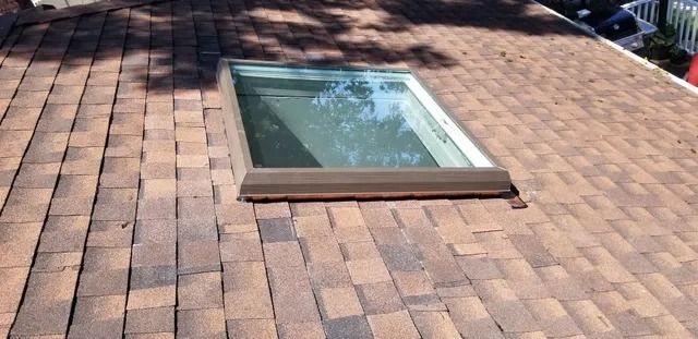 Rooftop skylight set into a shingled roof under sunlight, with trees reflected in the glass.