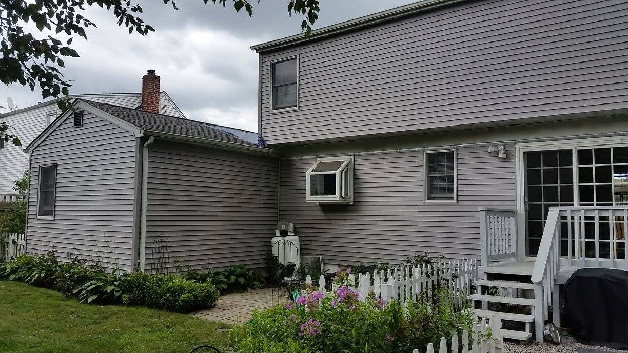 Gray house exterior with white trim, window air conditioner, and small garden under cloudy sky