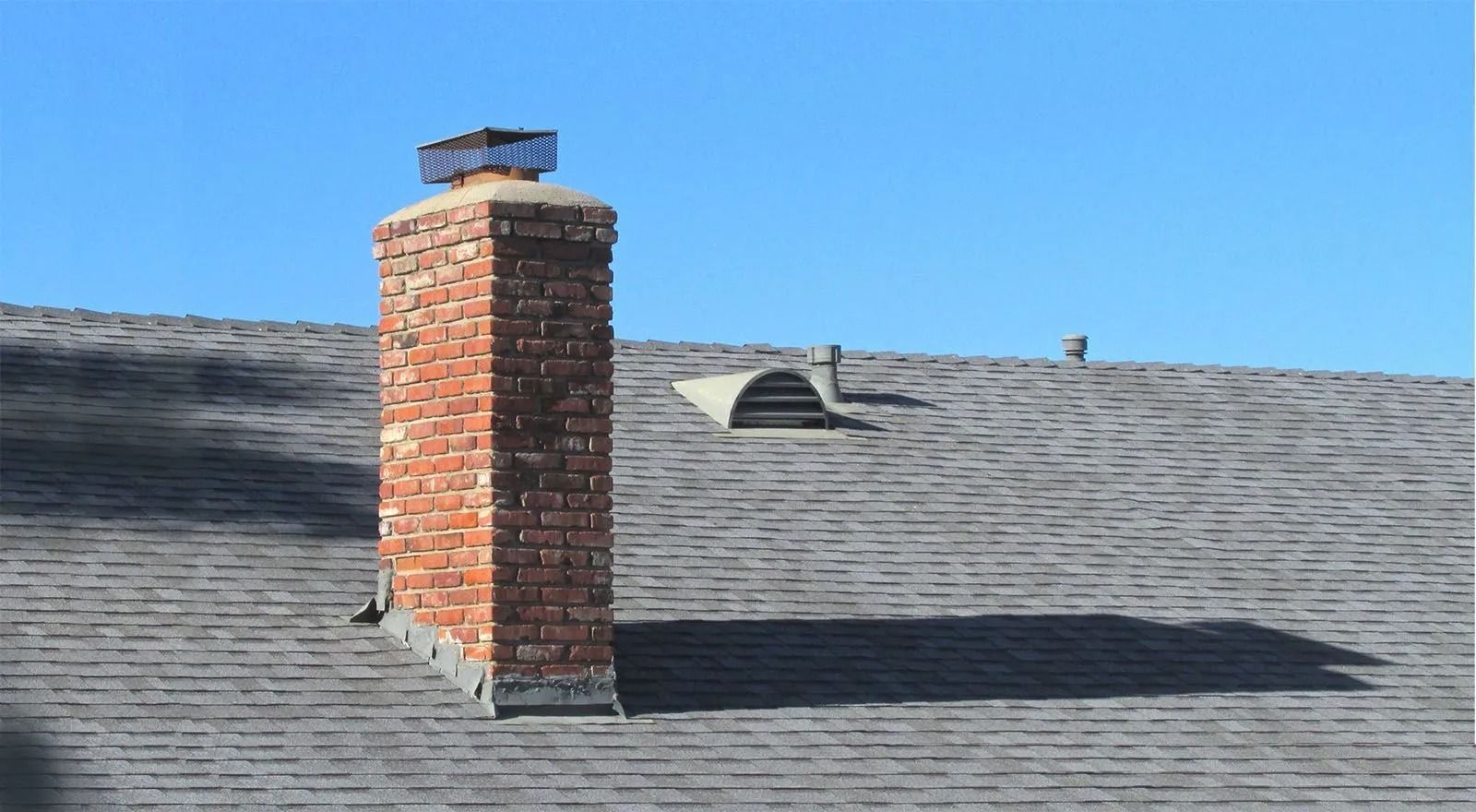 Brick chimney on a gray shingled roof under a clear blue sky