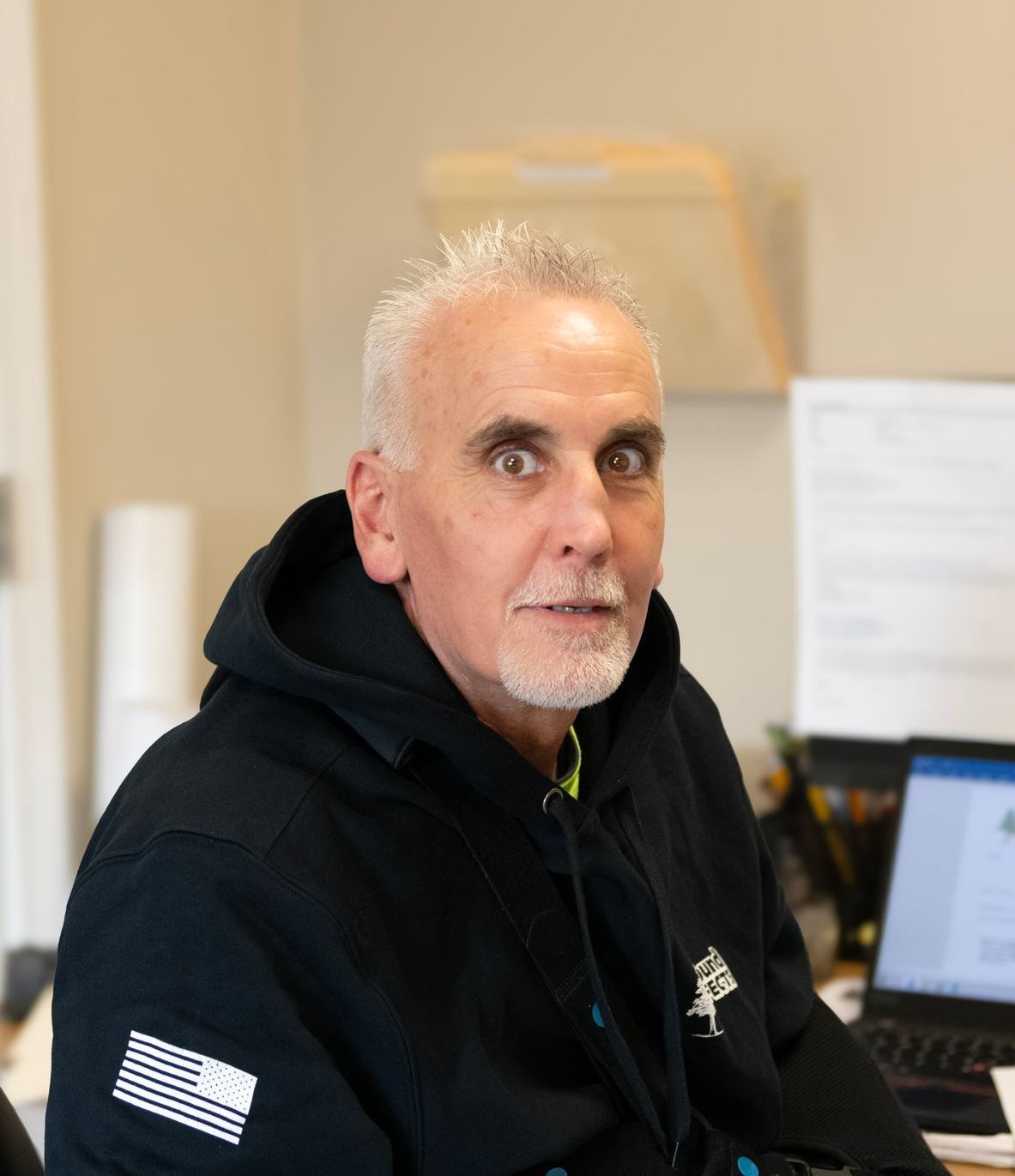 Man with gray hair and beard wearing a black hoodie looking surprised, sitting at a desk.