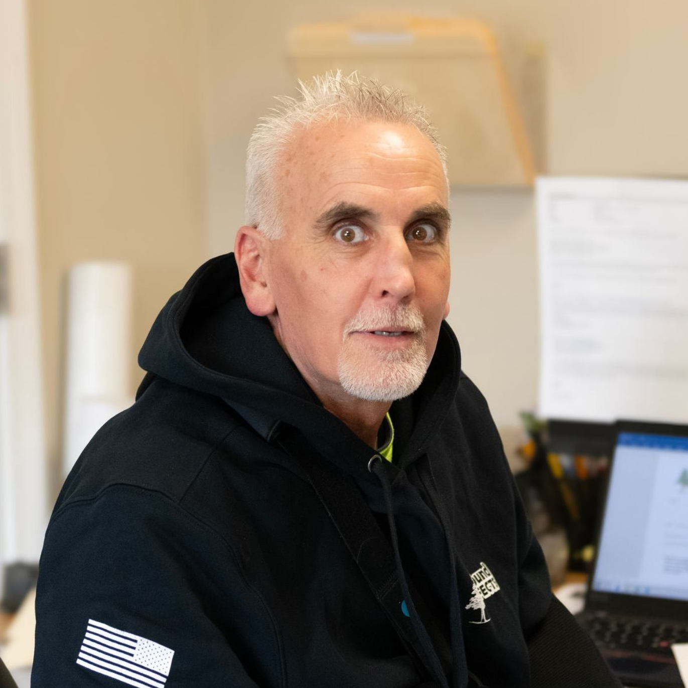 Man with gray hair and beard wearing a black hoodie looking surprised, sitting at a desk.