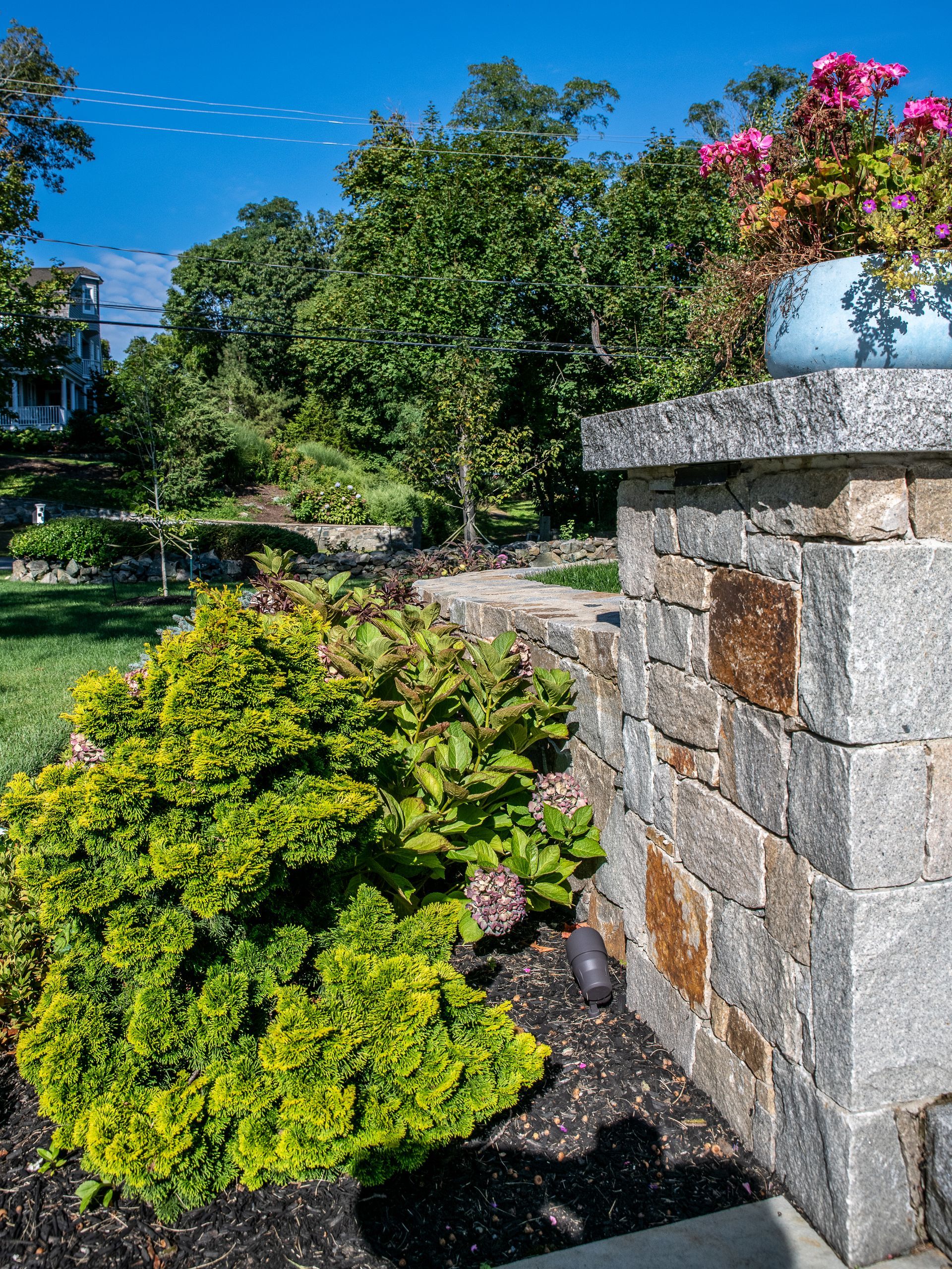 Stone pillar with blue flower pot, yellow-green shrub, trees, and lawn under a bright blue sky.