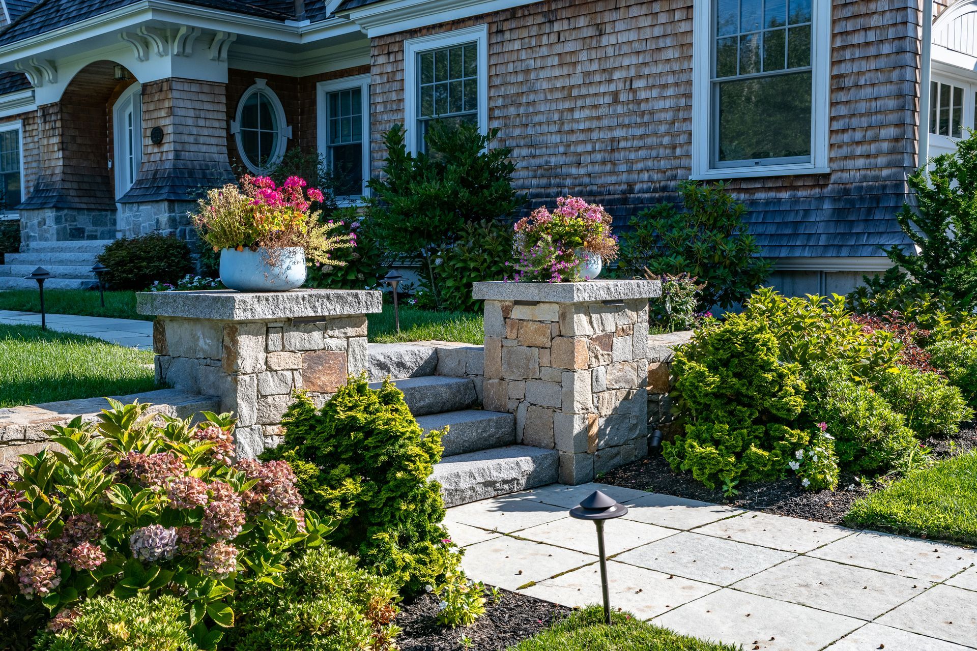 Stone pillars with flower pots flank steps leading to a brick house with a landscaped yard.