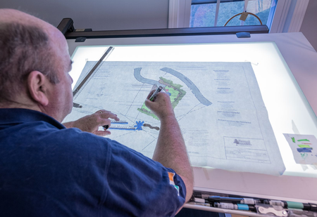 Man using a pen to draw on a lit architectural plan; studio setting.