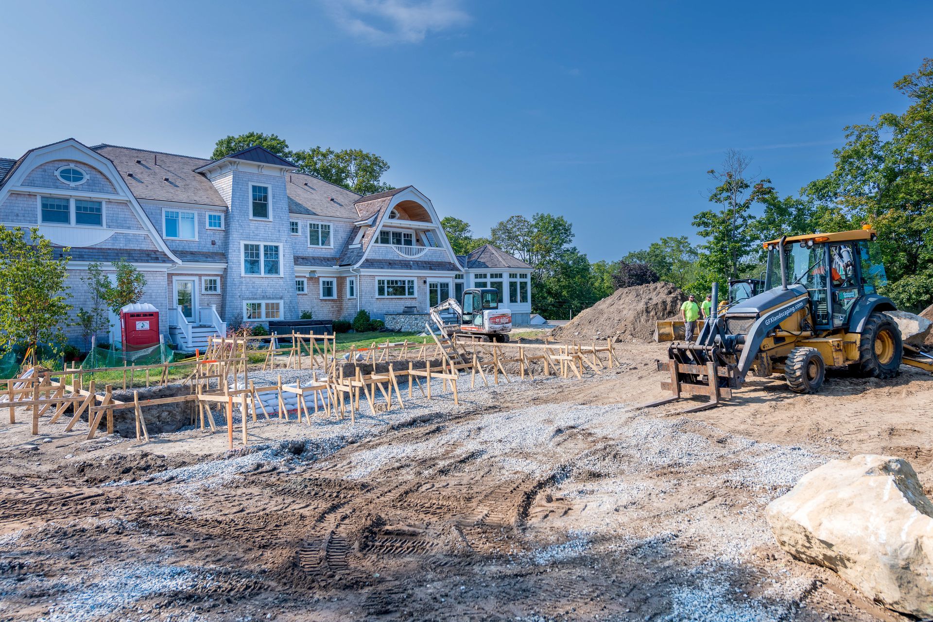 Construction site with a large house. A backhoe, and workers near lumber and dirt.