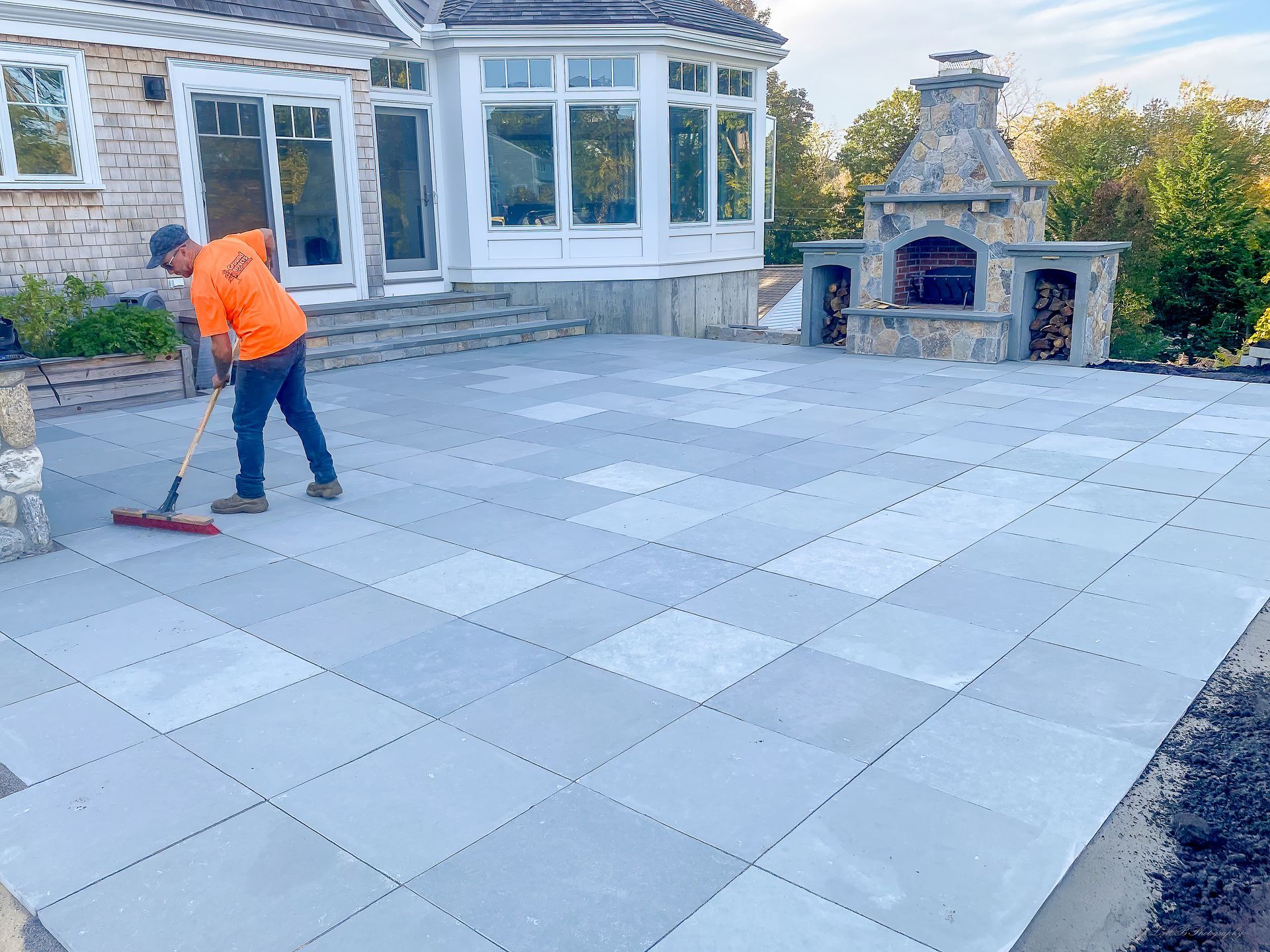 Man sweeping large, grey paver patio near a house with an outdoor fireplace.