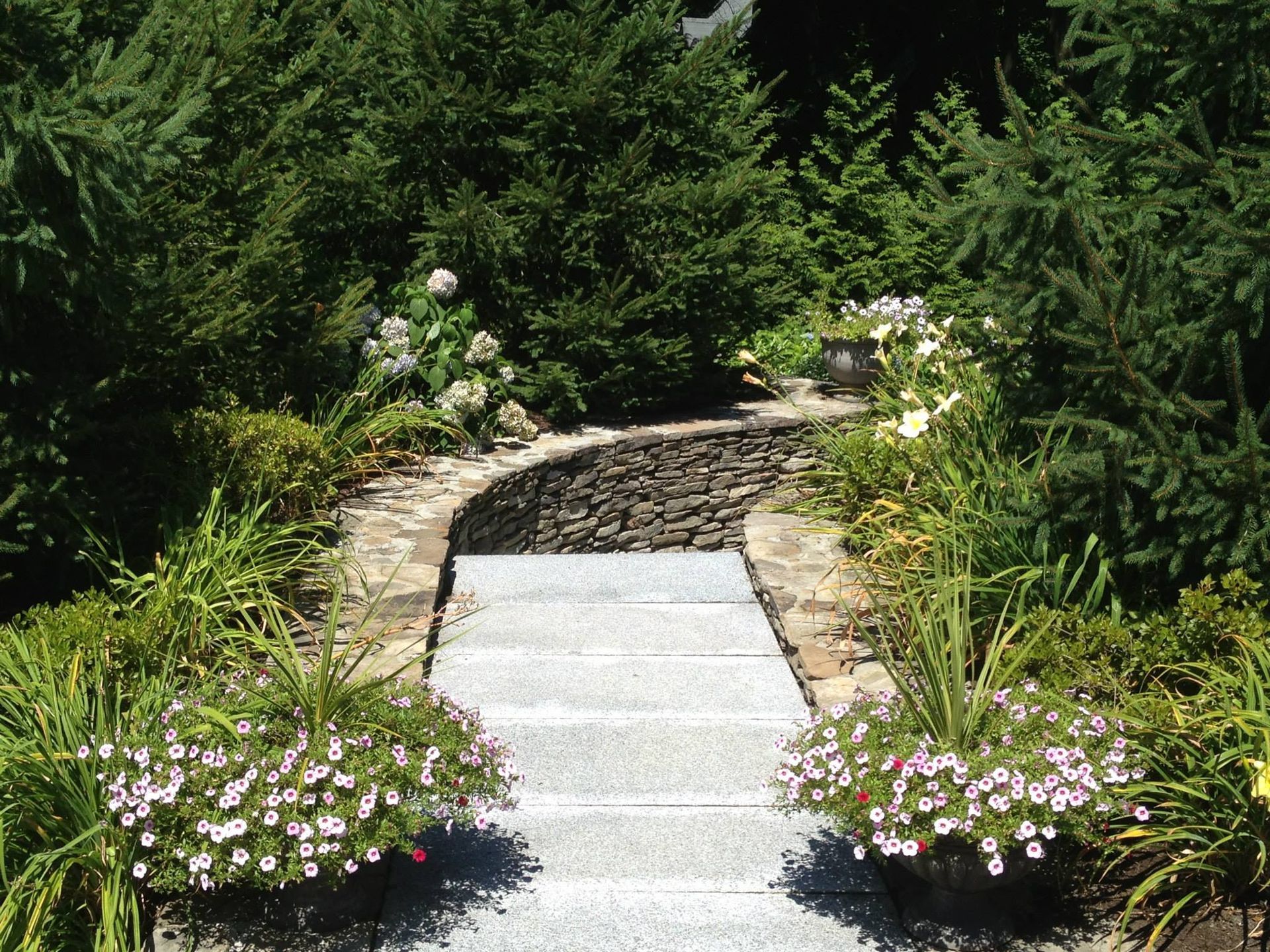 Stone pathway leading up to a small wall, surrounded by green trees, and flower pots on each side.