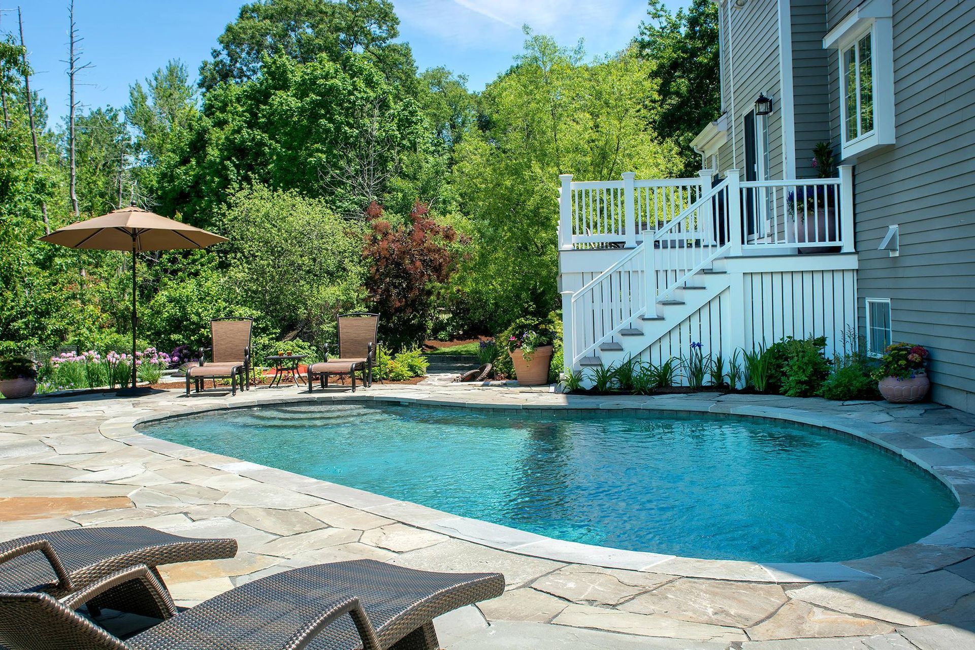 Pool surrounded by stone patio, lounge chairs, and lush greenery, with stairs leading to a deck on a house.