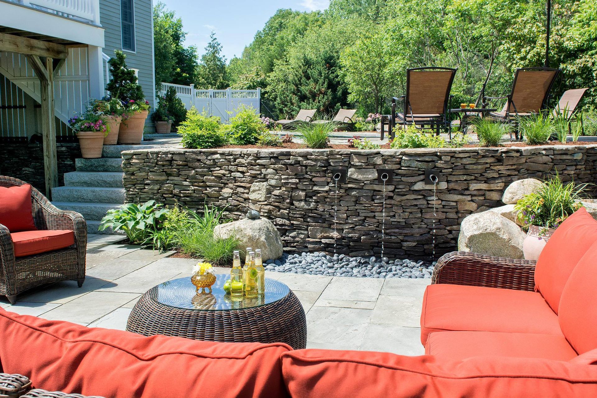 Patio with stone wall water feature, red sofa, wicker chairs, and greenery. Sunny outdoor setting.
