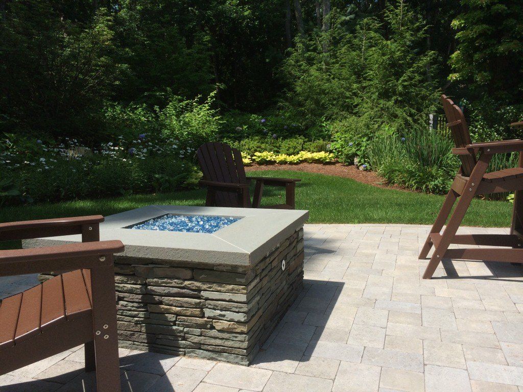 Stone fire pit on a patio with chairs; green trees and lawn in the background on a sunny day.