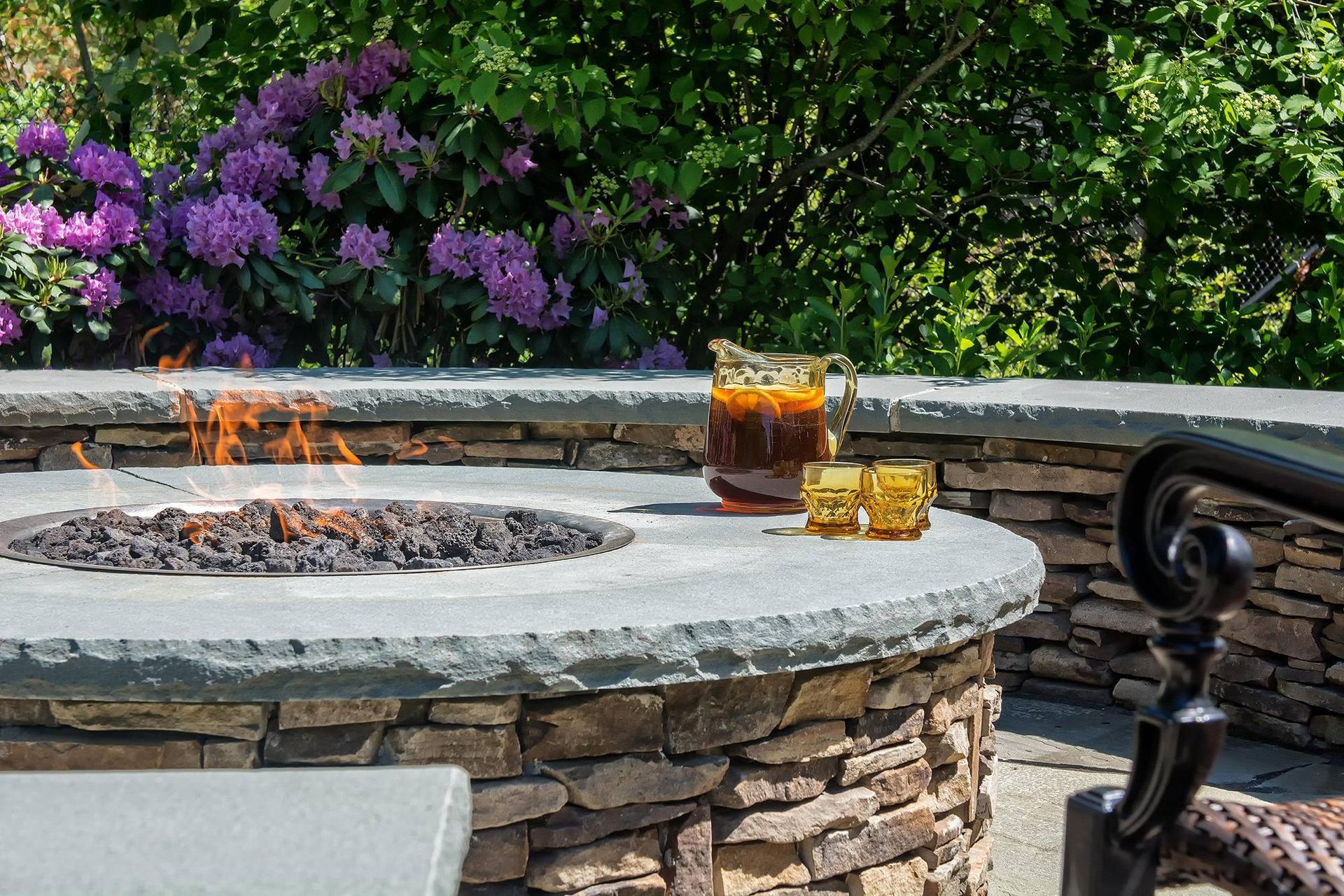 A stone fire pit with burning flames, a pitcher, and glasses sit on a stone wall with purple flowers in the background.