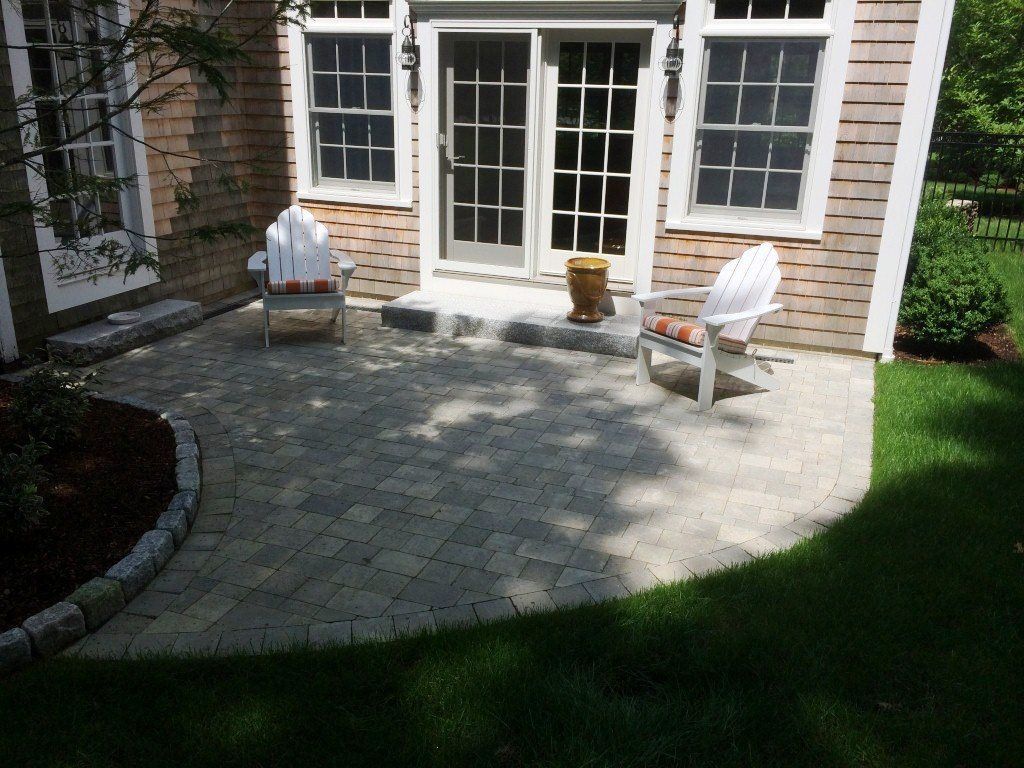 Paver patio with white chairs, next to a home with sliding glass doors, surrounded by green grass.