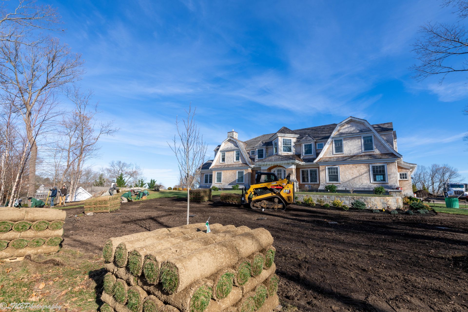 A house under construction with a yellow bulldozer and rolls of sod on a sunny day.