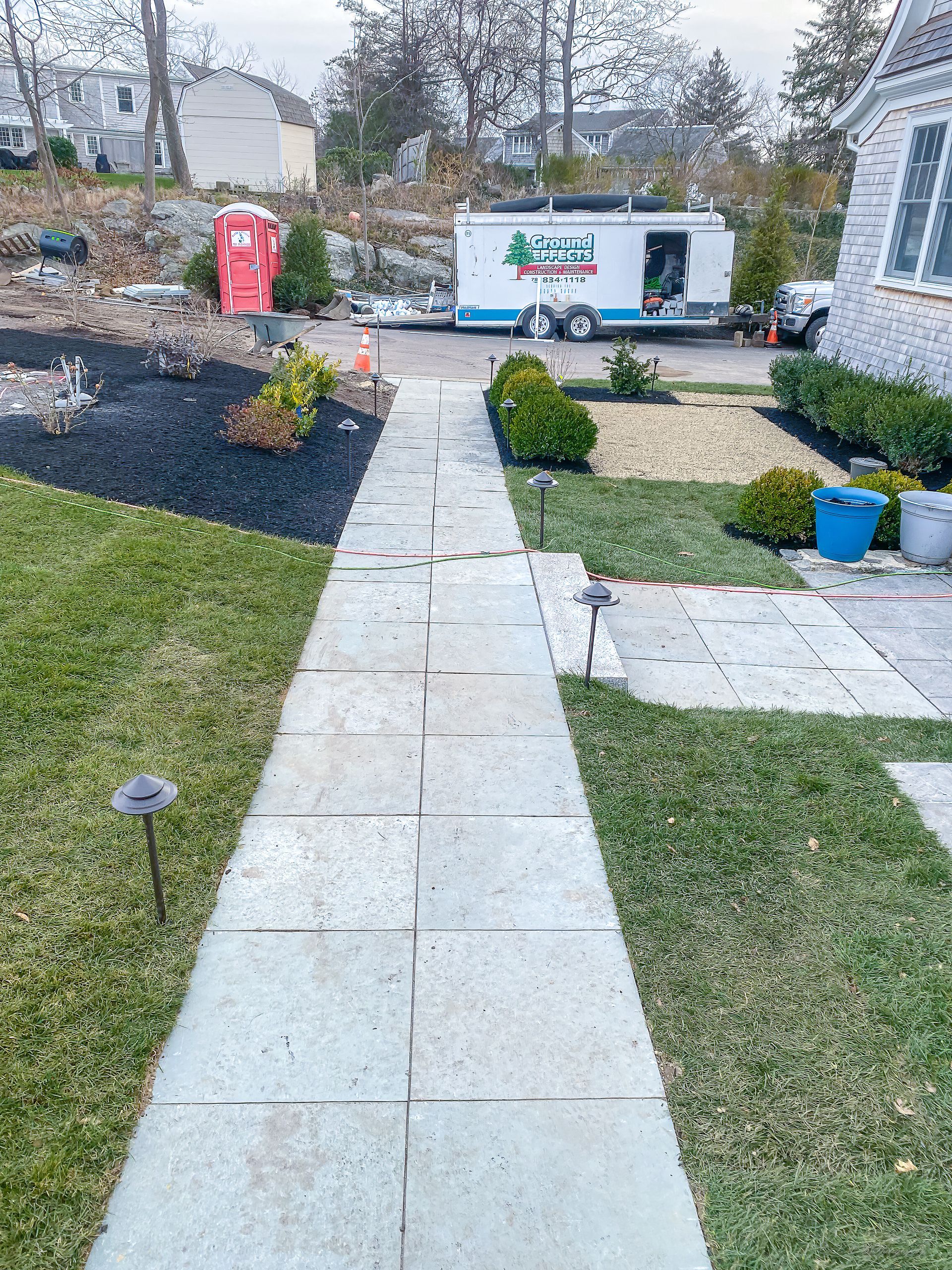 Concrete walkway leading to a trailer with landscaping work in progress; red portable toilet visible.