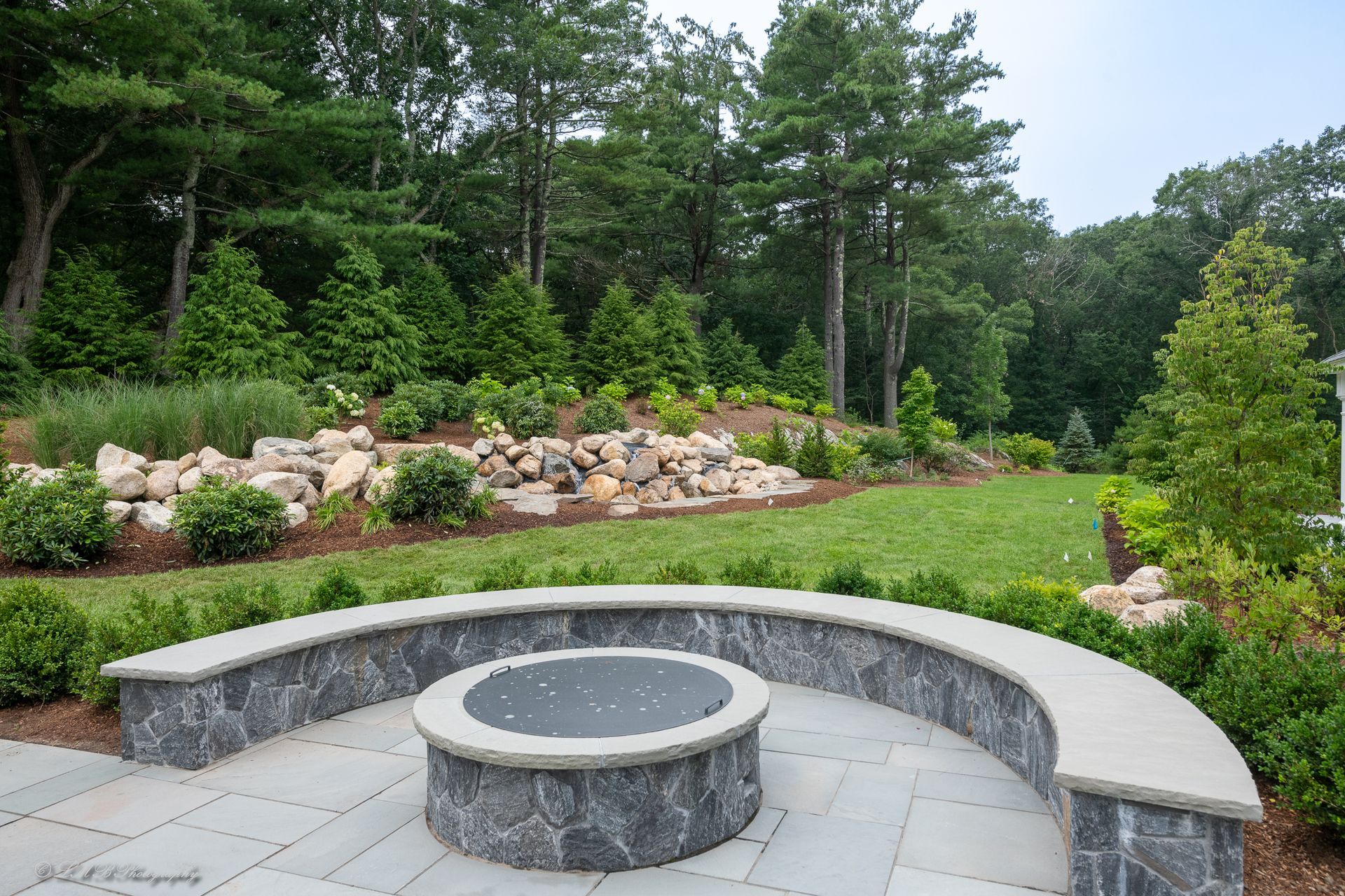 Outdoor patio with fire pit and curved seating. Green lawn and trees in the background.