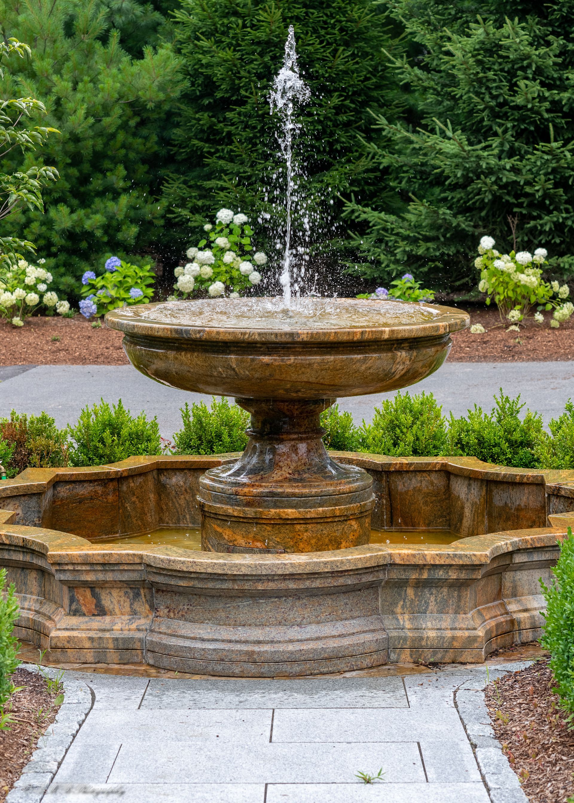 Stone fountain with water spraying into the air, surrounded by greenery and a stone walkway.