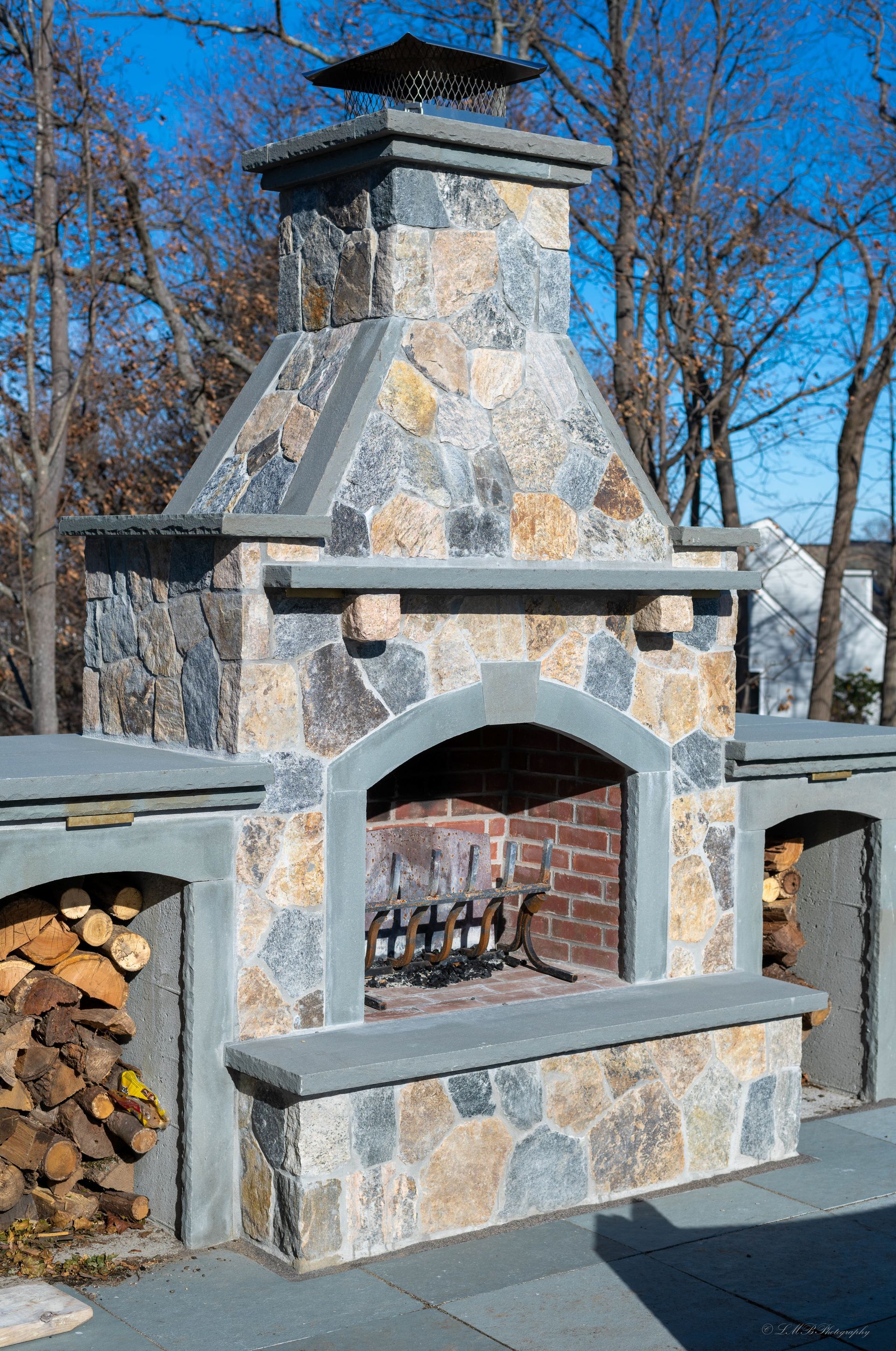 Stone outdoor fireplace with brick interior and wood storage on either side, blue sky.