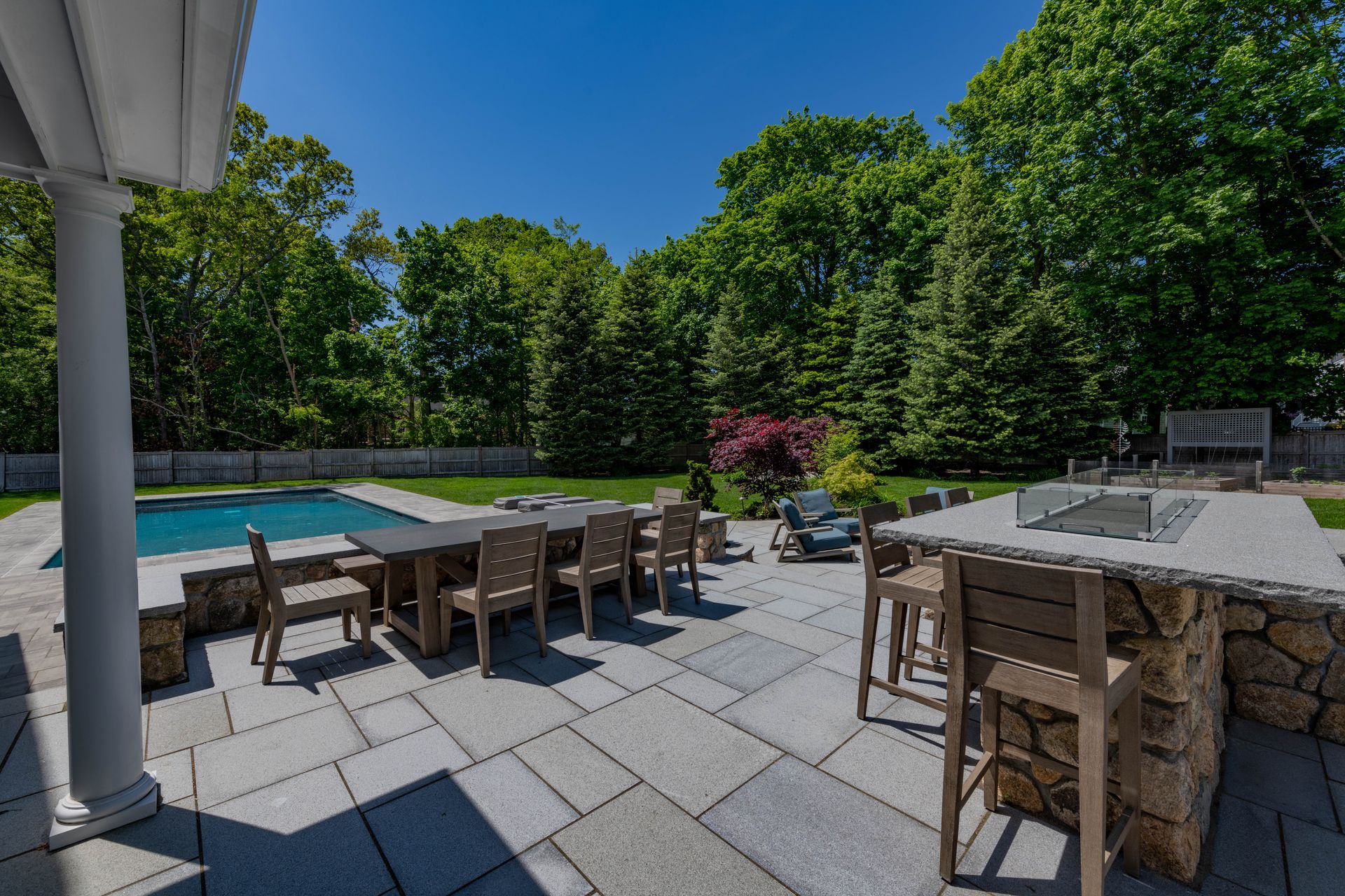 Patio with outdoor dining table, grill, and pool. Trees and blue sky in background.