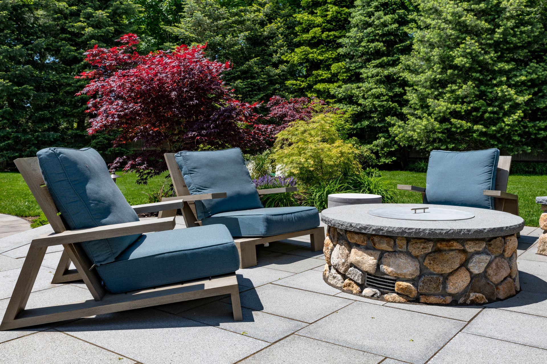 Patio with modern blue chairs around a stone fire pit, lush green and red foliage in the background.