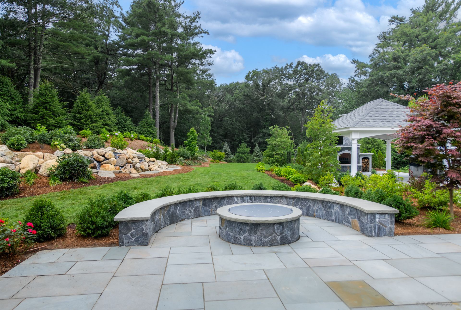 Stone patio with fire pit, curved stone seating, green lawn, trees, and gazebo under a blue sky.