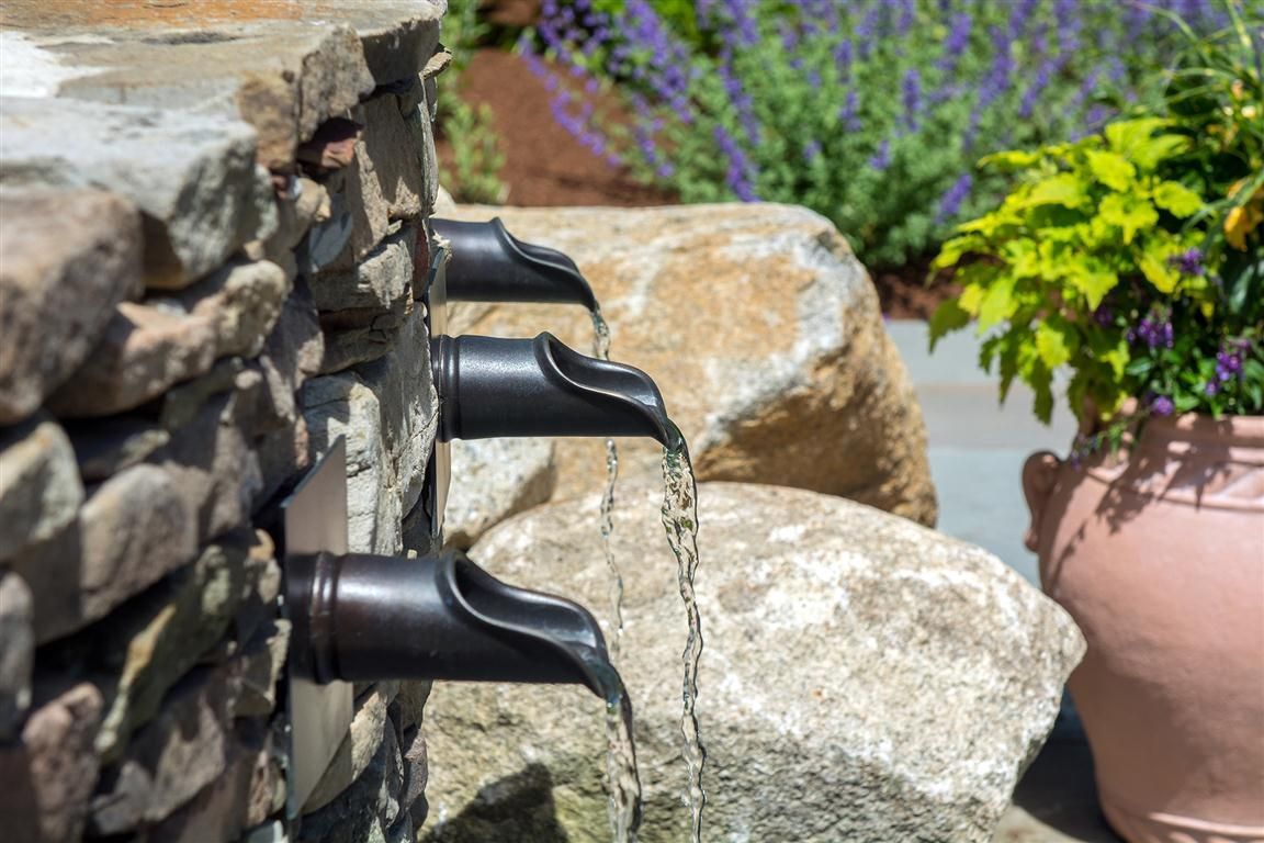 Water fountain with three spouts cascading water, stone wall and rocks, green plants in background.