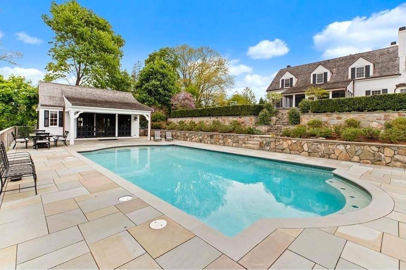 Swimming pool and pool house with a large white house in the background. Blue sky and stone patio.