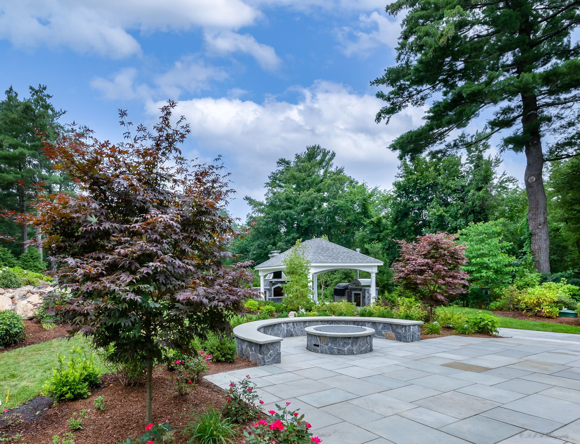 Backyard patio with fire pit, gazebo, and surrounding landscaping under a partly cloudy sky.
