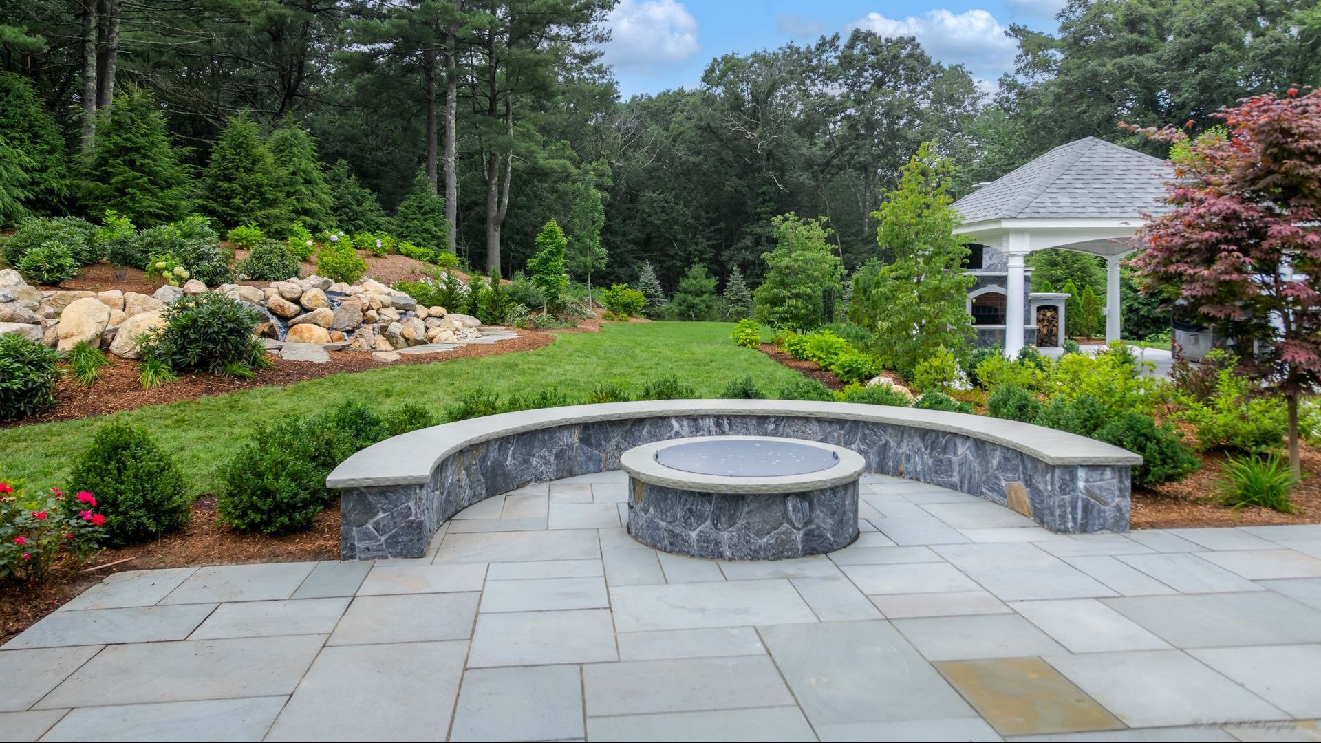 Stone patio with a fire pit and curved seating, surrounded by landscaping, with a gazebo in the background.