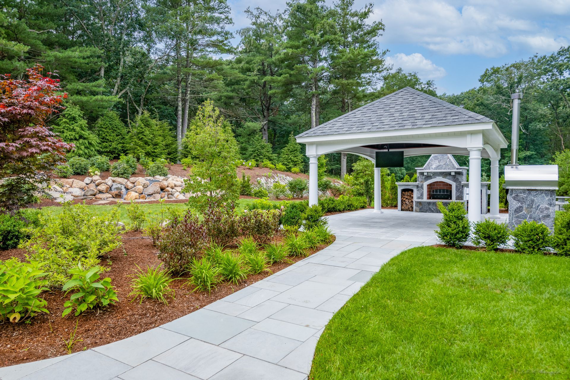Stone patio with gazebo, outdoor kitchen, lush landscaping, and trees.