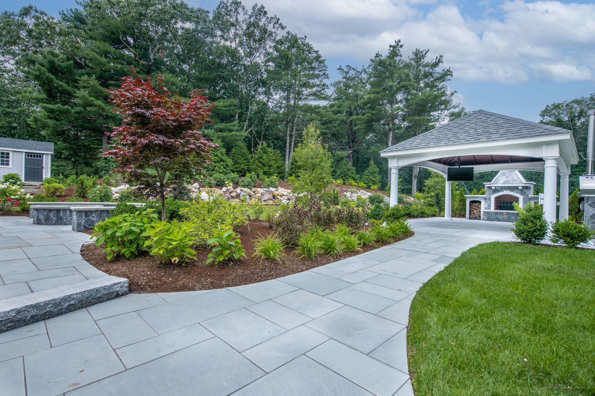Paver patio with a gazebo, outdoor fireplace, and garden with trees.