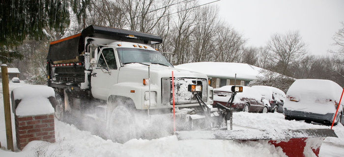 Snowplow clearing snow from a residential driveway. The truck is white with an orange dump bed, and the plow is red.