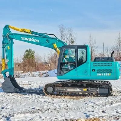 Turquoise Sunward excavator on snowy ground beside bare trees under a blue sky