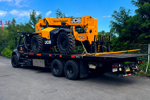 Orange JCB telehandler loaded on a black flatbed trailer on a roadside under a blue sky