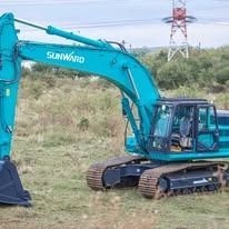 Turquoise SUNWARD excavator on tracks in a grassy field near power lines