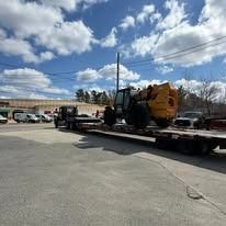 Flatbed trailer hauling a yellow construction machine in a parking lot under a cloudy sky