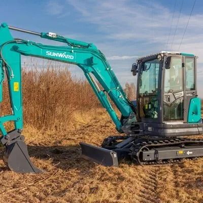 Turquoise excavator in a muddy field, with tall dry grass and a cab on the right.