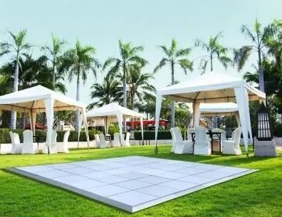 Outdoor event setup with white tents, chairs, and a dance floor on a green lawn palm trees.