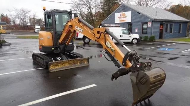 Orange backhoe parked on a wet street beside a gray building with a white truck nearby