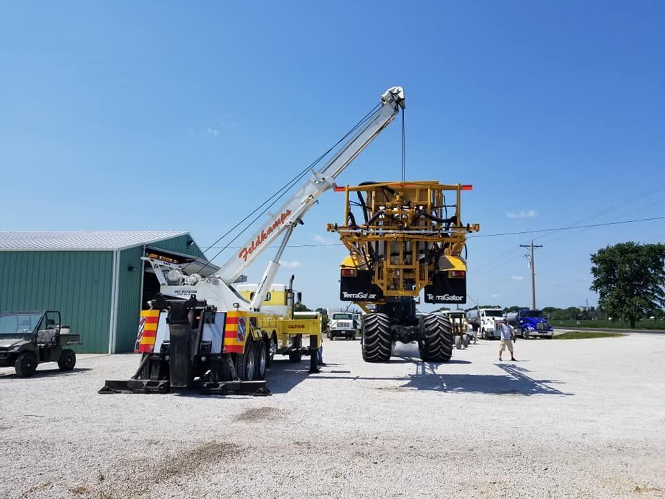 A large tow truck lifting a yellow agricultural machine in a gravel lot on a sunny day.