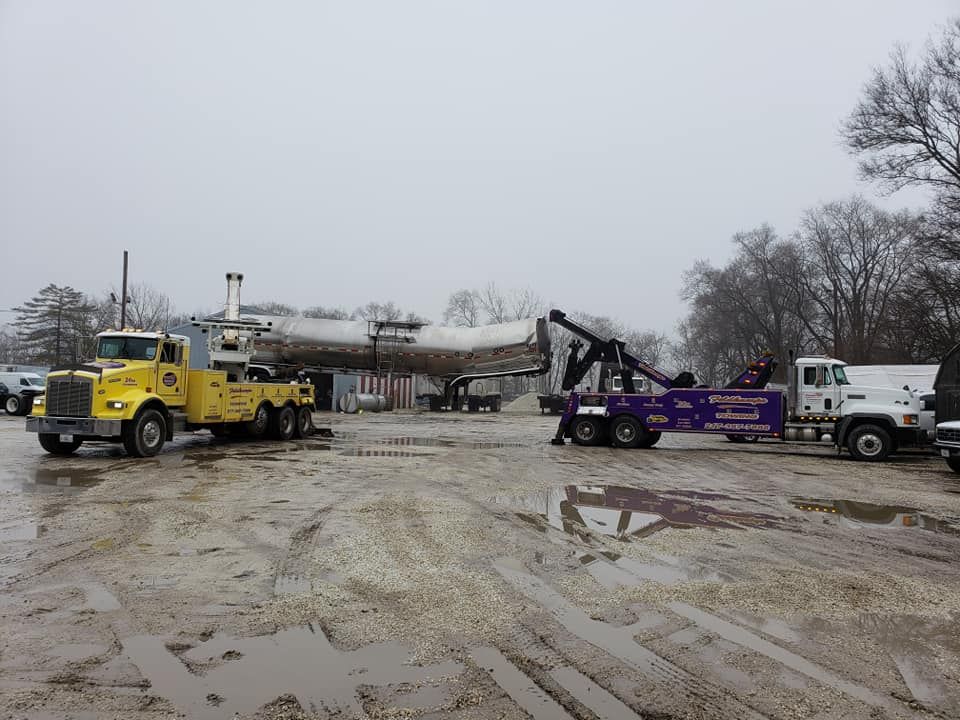 Three tow trucks surrounding an airplane fuselage on a muddy lot; overcast sky.