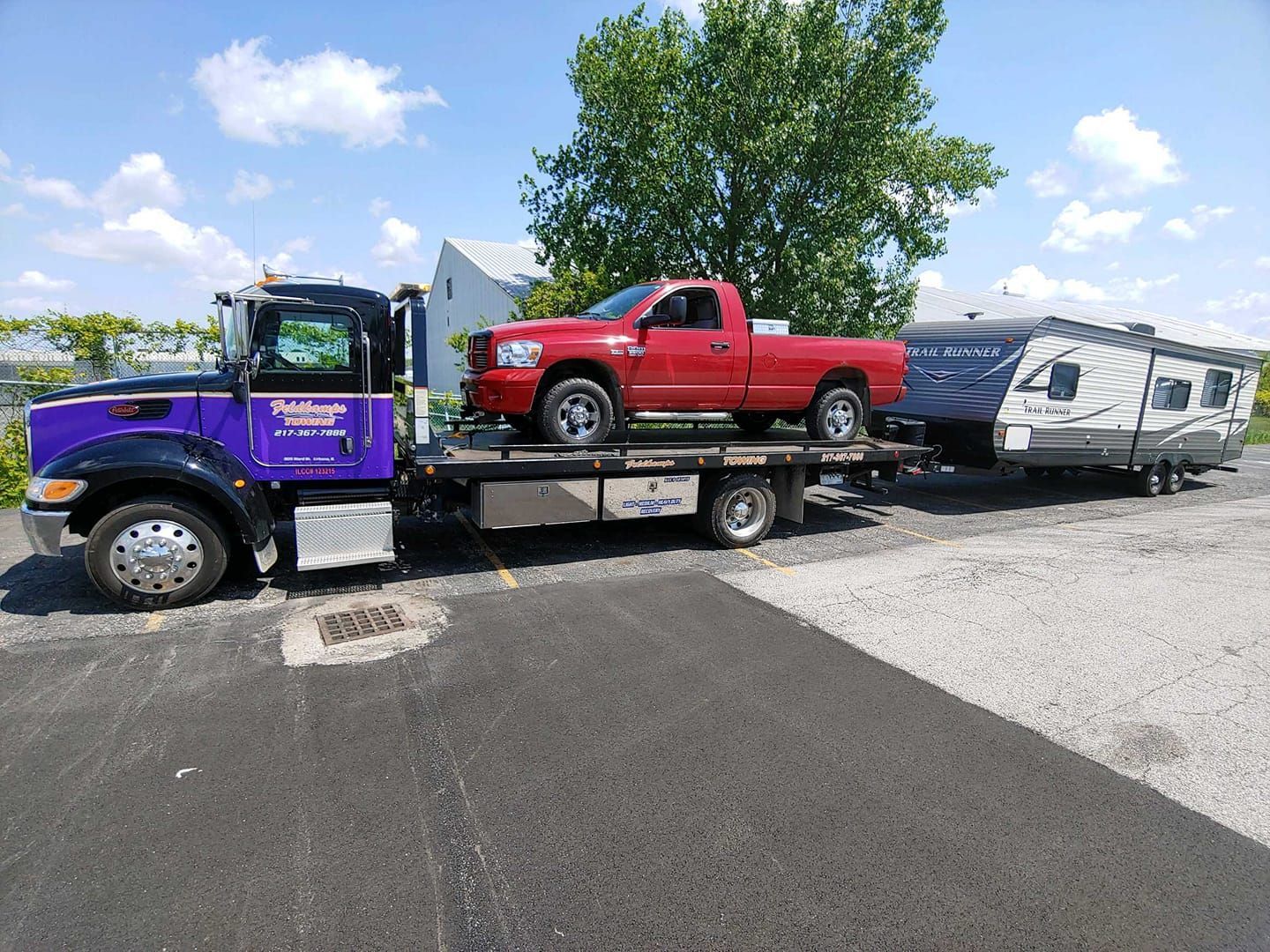Purple tow truck transporting a red pickup truck and a travel trailer on a paved lot under a blue sky.