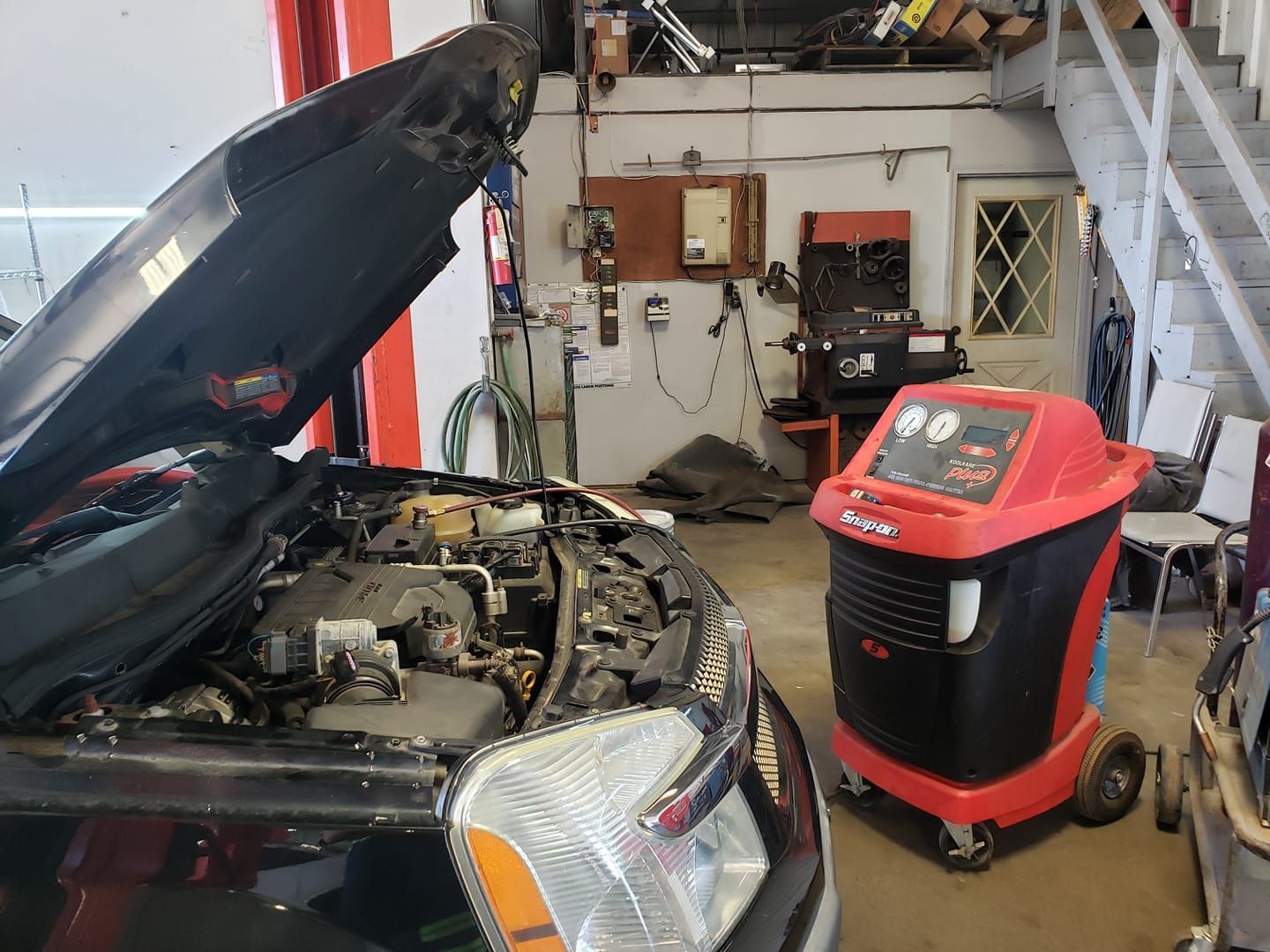 Car with open hood in auto shop next to a red/black AC recharge machine.