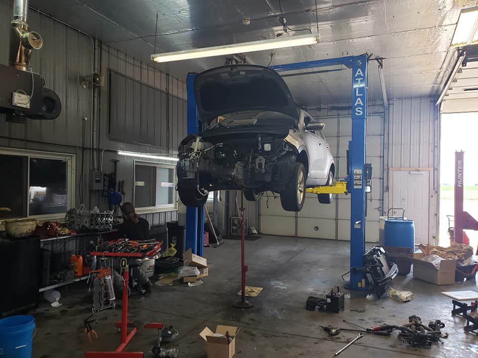 Car on a lift in a garage with open hood. Tools and parts on the floor, repair in progress.
