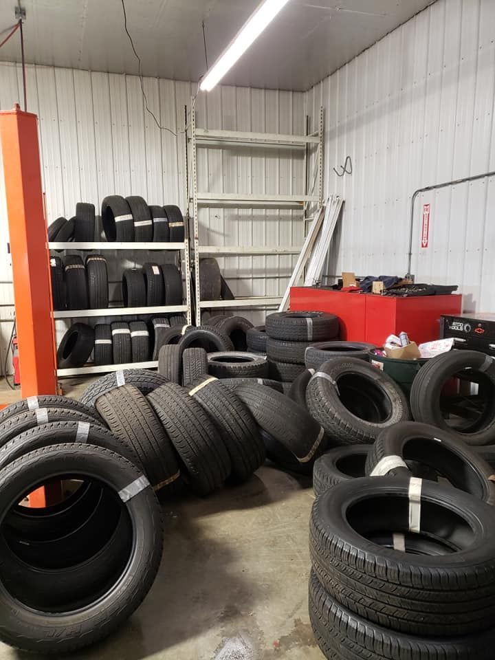 Tires stacked in a cluttered garage, some on shelves, others in piles. A red machine and metal shelving are also visible.