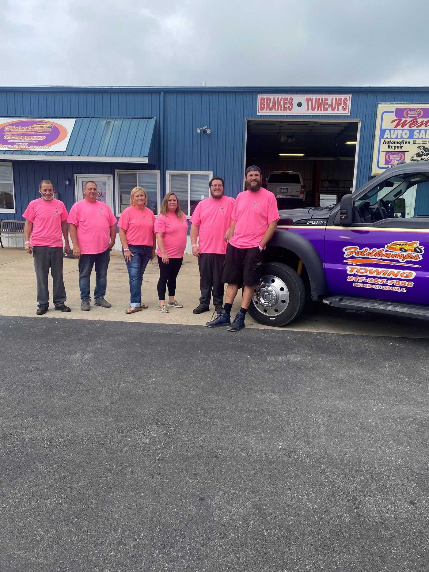 Group of people wearing pink shirts posing by a purple truck in front of a blue building with 