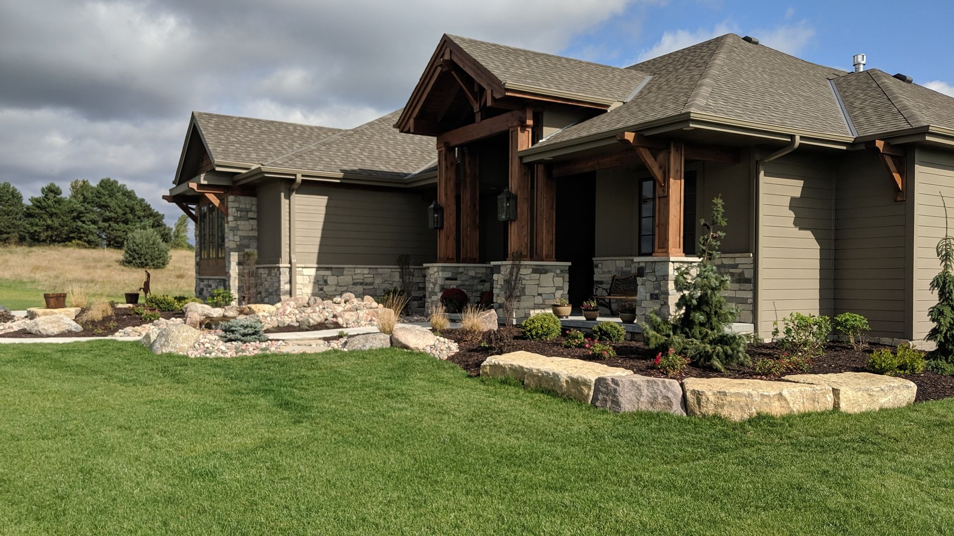 Beige house with stone accents, brown wooden supports, and landscaping against a cloudy blue sky.