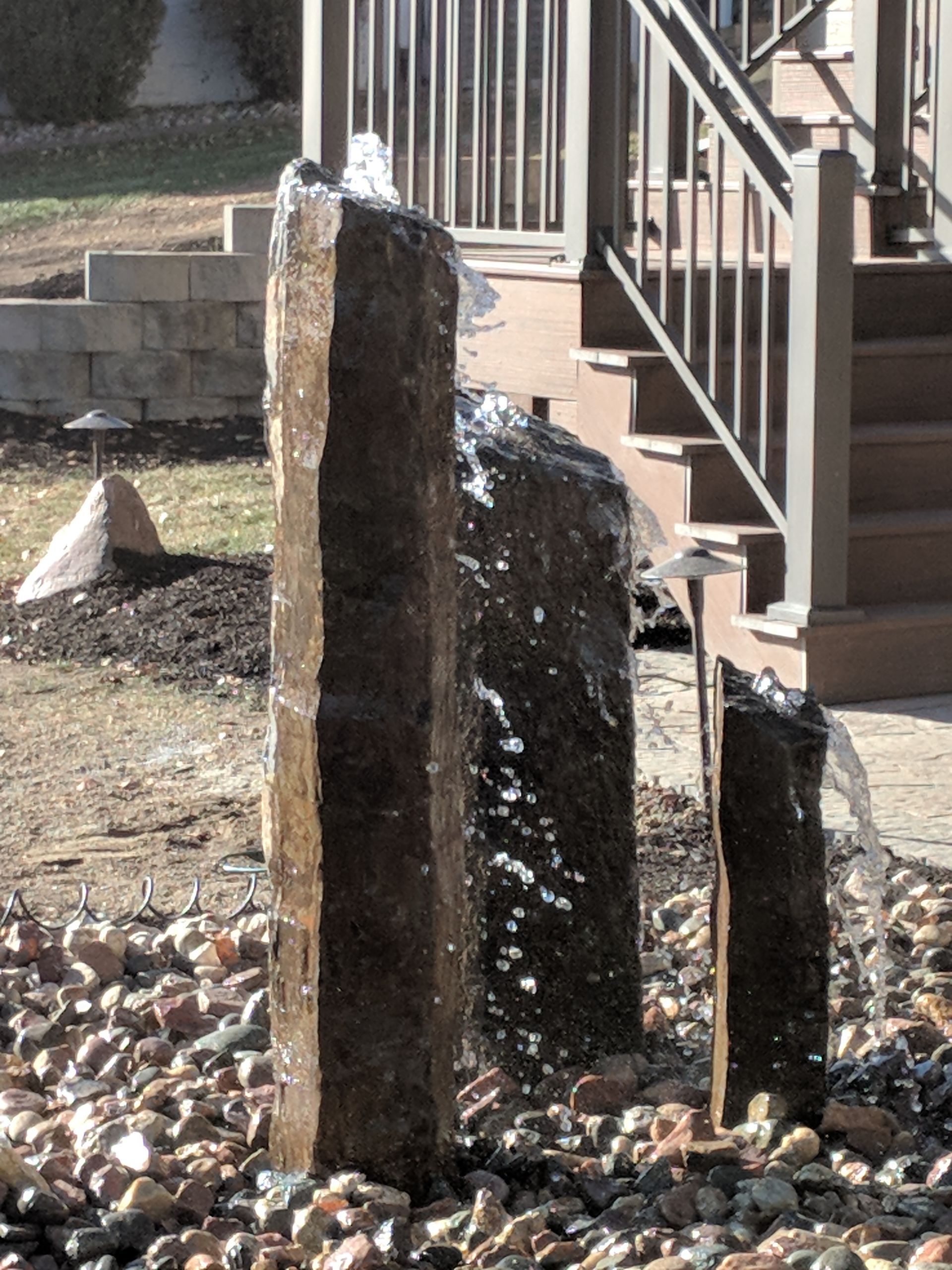 A three-tiered stone fountain with water cascading into a bed of rocks, near a wooden staircase.