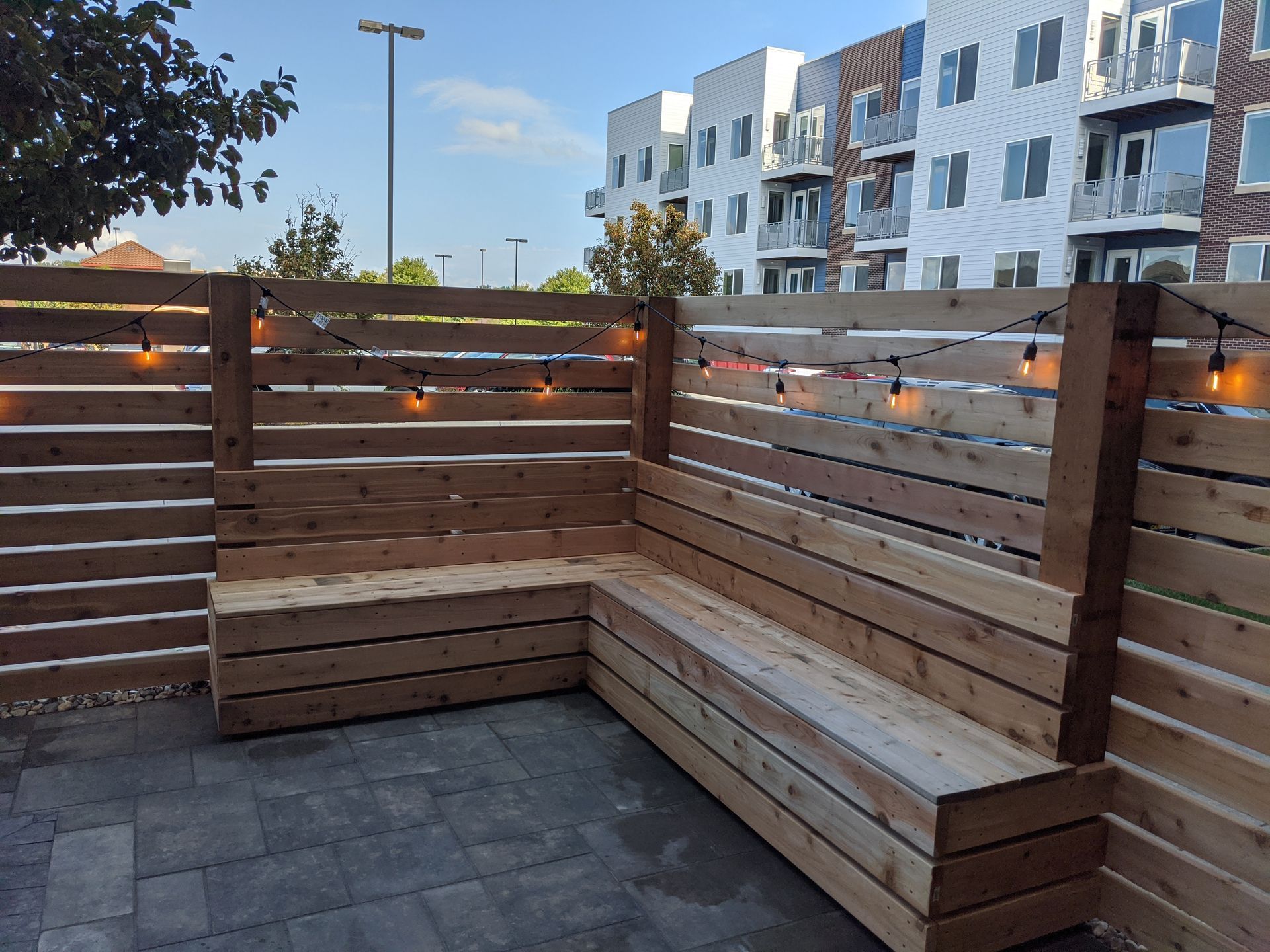 Wooden outdoor bench and fence, lit by string lights, on a patio, with a building in the background.
